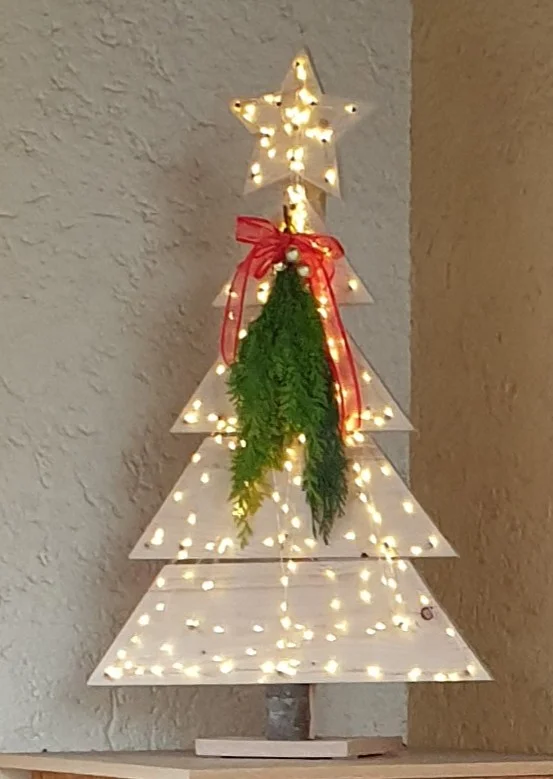 Dekorativer Weihnachtsbaum aus Holz mit Lichterkette und roter Schleife.