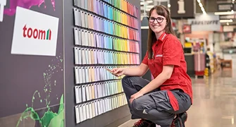 Eine Person in Arbeitskleidung kniet vor einer Wand mit vielen bunten Farbmustern.
