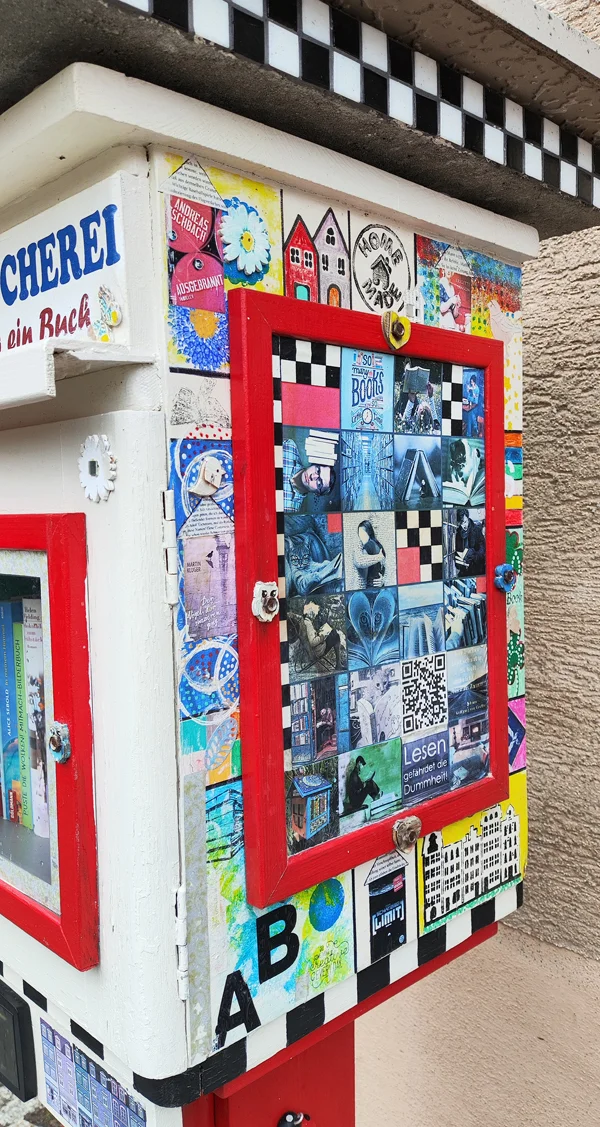 Ein bunt verzierter öffentlicher Bücherschrank mit roten Rahmen und vielen Collagen.