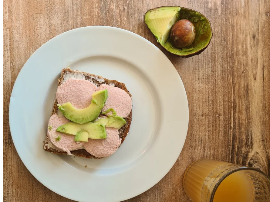 Draufsicht auf ein belegtes Brot mit Wurst und Avocado neben einem Glas Saft.
