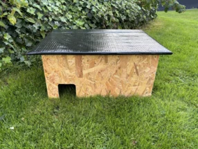 Ein Igelhaus aus Holzplatten mit schwarzem Dach steht auf einer Wiese.