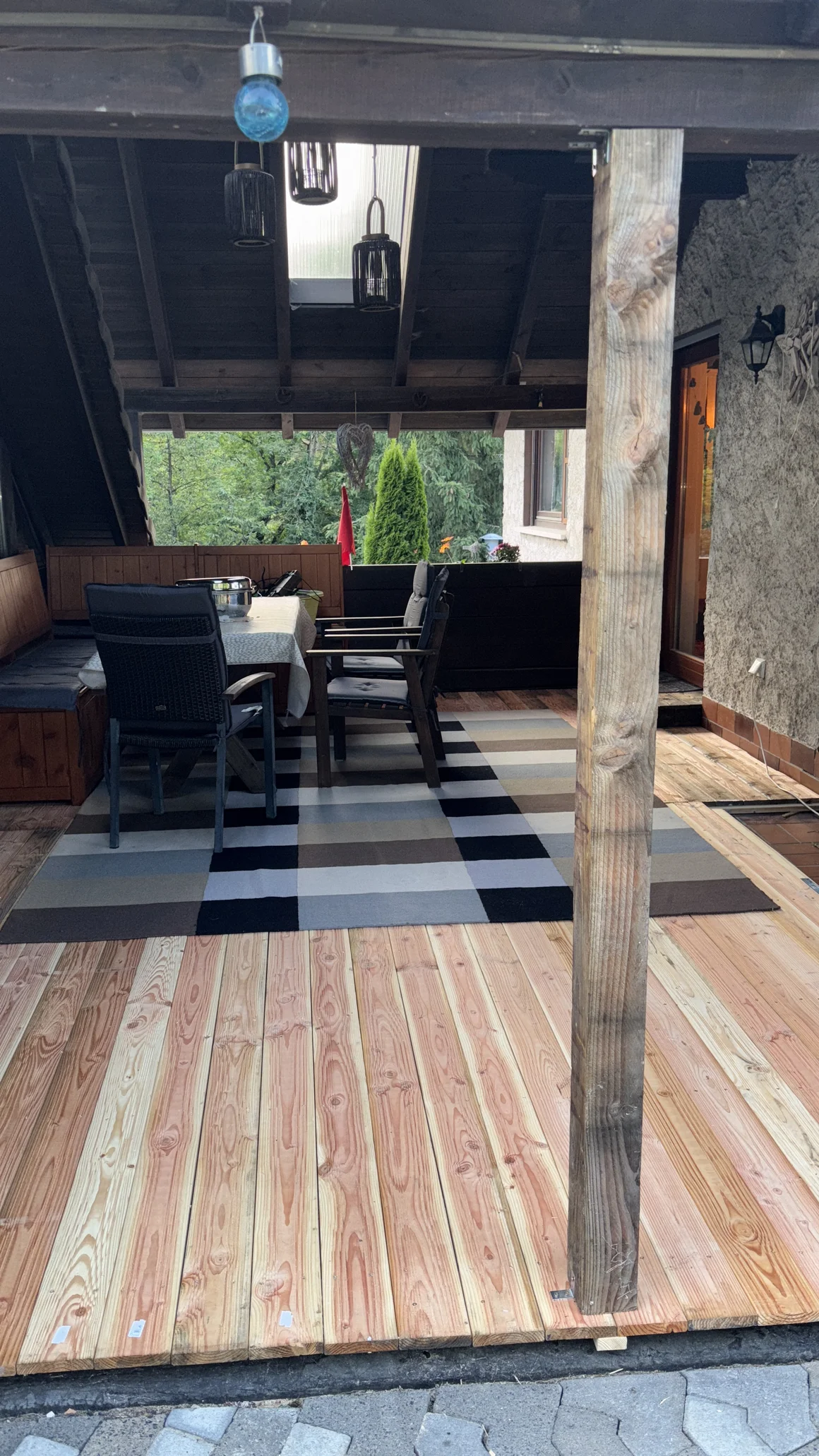 Eine seitliche Aufnahme einer überdachten Holzterrasse mit Gartenmöbeln und einem gestreiften Teppich.