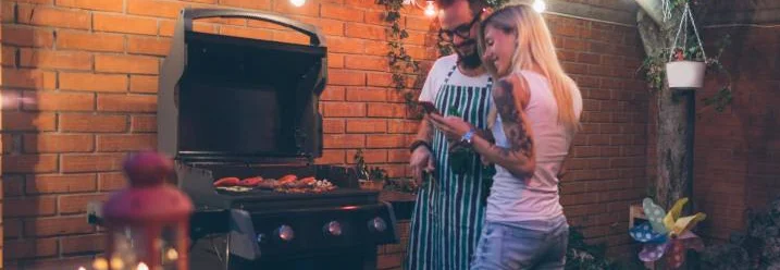 Zwei Personen schauen auf ein Smartphone und stehen vor einem Gasgrill.