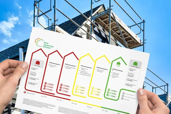 Hände halten einen Sanierungsplan vor einem Haus mit Baugerüst