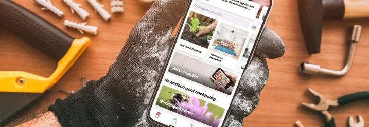 Hand hält Smartphone mit Baumarkt-App auf einem Tisch mit Werkzeug.