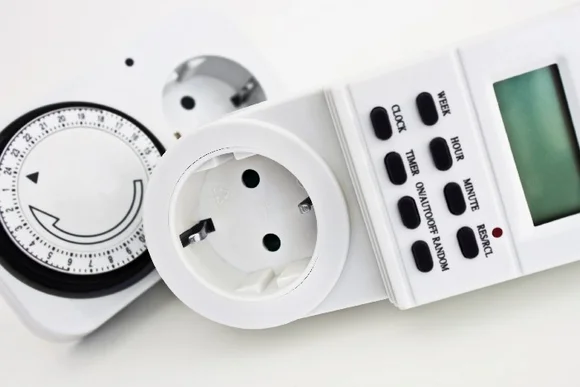 Eine digitale Zeitschaltuhr mit integrierter Steckdose und ein mechanischer Timer liegen auf einem Tisch.